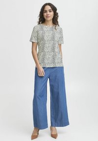 Top a maniche corte color crema con un motivo maculato nero, abbinato a pantaloni in denim blu a gamba larga e scarpe con tacco color nocciola.