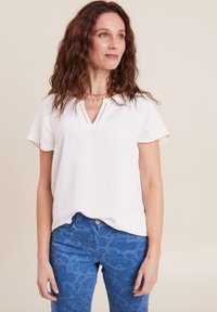 Weißes T-Shirt mit kurzen Ärmeln und V-Ausschnitt aus strukturiertem Stoff, kombiniert mit blauen Jeans mit Paisleymuster und Knopfverschluss.