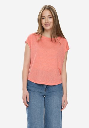 Ung kvinde smilende, iført en koral kortærmet top og blå jeans, stående foran en ensfarvet hvid baggrund.