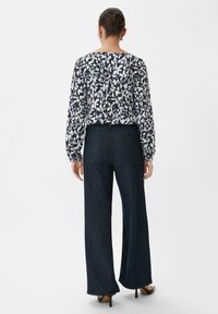 Blouse met lange mouwen in een marineblauw en wit abstract bloemenpatroon, gecombineerd met een hoge taille donkere denim wijde broek en zwarte hakken.