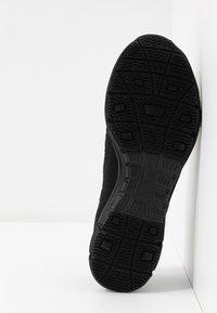 Semelle de chaussure de sport noire avec un motif de bande de roulement texturé, appuyée contre un mur et une surface blancs.