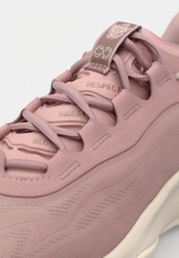 Calzado rosa con superficie texturizada, sistema de cordones y texto en relieve. Material suave con una forma elegante y redondeada y suela en contraste.
