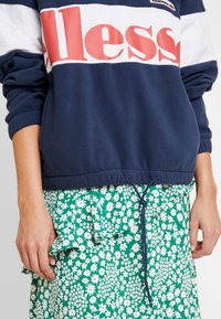 Marinblå fleecepullover med vit och röd rand, med "Aless"-logotyp. I kombination med en grön blommig volangkjol med vita prästkragar.