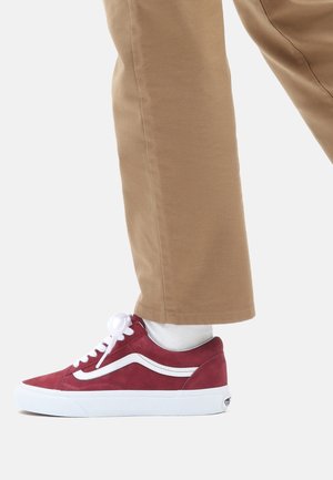 Persona che indossa pantaloni beige e calze bianche, che mostra una sneaker rossa bassa con lacci bianchi e un dettaglio a striscia bianca.