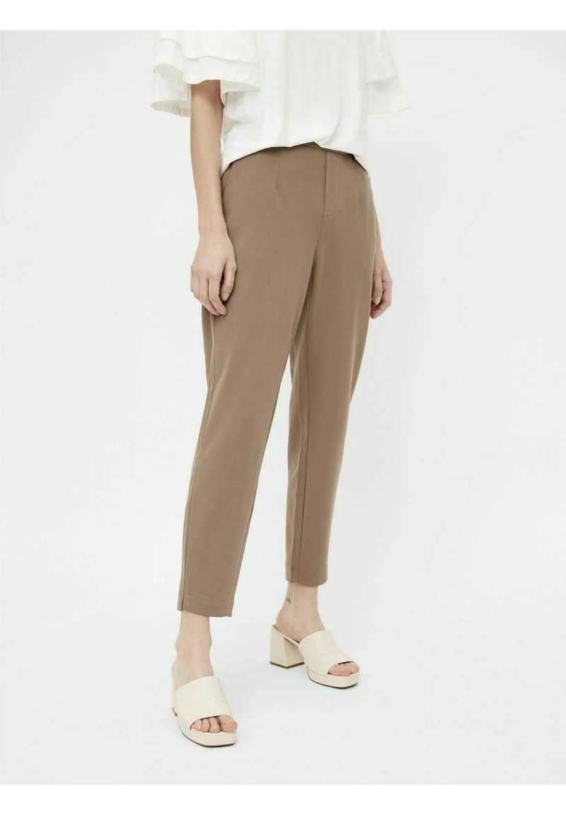 Femme portant un pantalon beige tailleur longueur cheville, une blouse blanche à manches courtes et des sandales beiges à talon bloc ouvrant les orteils, debout sur fond blanc.