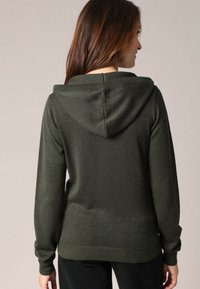 Donkergroene hoodie gemaakt van textuurstof. Heeft een geribde onderkant en boorden, met een subtiele glans. Geen opvallende patronen of accenten.