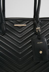 Sac à main en cuir noir avec couture chevron, plaque logo Aldo dorée, bandoulière réglable avec boucle dorée et étiquette bagage Aldo attachée.