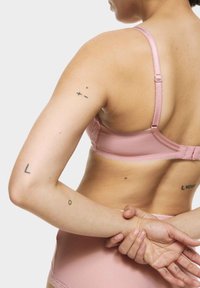 Ljust rosa bh med justerbara remmar, spetskanter och en mjuk yta. Tatueringar synliga på armarna, med ett fokuserat perspektiv på ryggen.