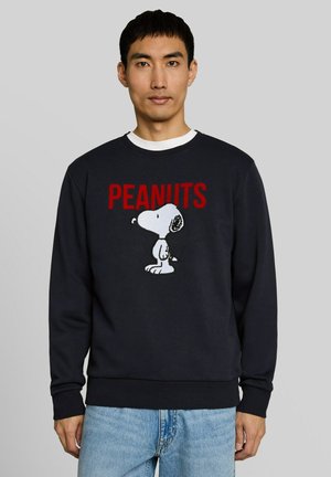 Man draagt een zwarte trui met rode tekst "PEANUTS" en witte Snoopy-afbeelding, gecombineerd met lichtblauwe spijkerbroek, staand tegen een grijze achtergrond.