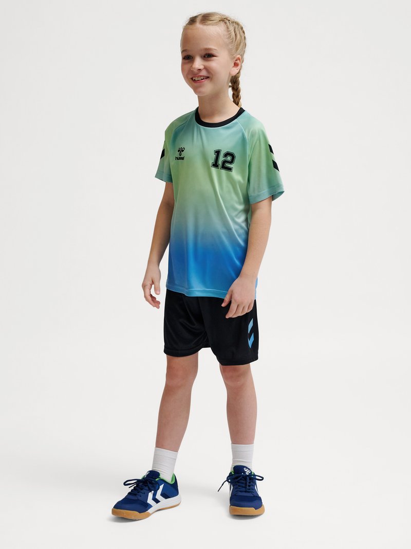 Junges Mädchen mit geflochtenem Haar, das ein Sporttrikot mit grün-blauem Farbverlauf und der Nummer 12 trägt, schwarze Shorts, weiße Socken und blaue Sportschuhe.