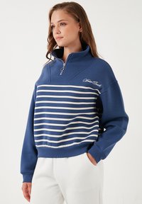 Maglione blu con strisce orizzontali bianche, colletto con mezza zip e polsini a costine. Presenta un dettaglio ricamato "Venice Beach".