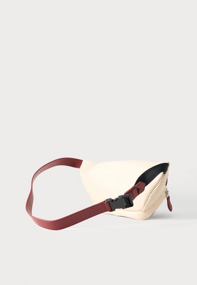 Συμπαγής μπεζ τσαντάκι χιαστί με διάτρητη πλευρική λεπτομέρεια, βυσσινί ρυθμιζόμενο ιμάντα και μαύρο πλαστικό κούμπωμα σε μονόχρωμο φόντο.