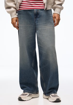 Personne portant un jean bleu délavé à jambes larges, des baskets grises et blanches, une chemise rayée rouge et blanche et une veste beige clair.