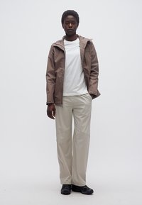 Άτομο που στέκεται φορώντας καφέ μπουφάν, λευκό μπλουζάκι, μπεζ παντελόνι και μαύρα παπούτσια, μπροστά σε απλό φωτεινό φόντο.