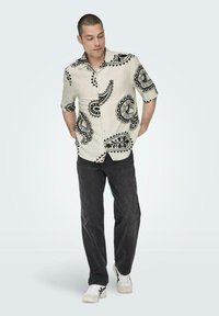 Beige Kurzarmshirt mit schwarzen Paisleymustern, kombiniert mit dunklen Jeans und weißen Sneakers. Verfügt über ein knöpfbares Design und einen lockeren Schnitt.