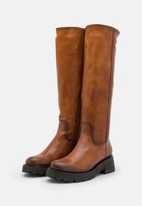 Une paire de bottes en cuir brun hautes avec des semelles à crampons noires, des bouts arrondis et un minimum de détails de couture sur un fond blanc uni.