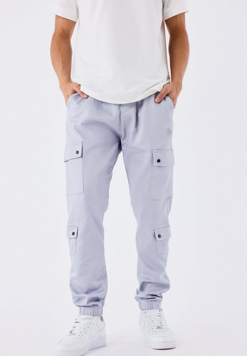 Uomo che indossa pantaloni cargo viola chiaro con polsini elastici, sneakers bianche e una maglietta bianca, in piedi con le mani in tasca.