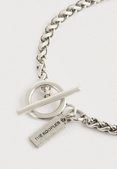 Pulsera de cadena de plata con cierre de palanca y placa rectangular grabada con "THE KOOPLES" sobre un fondo blanco.