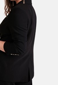 Zwarte blazer met een gestructureerde kraag en opgerolde mouwen, met een rugopening en gouden knoopaccenten op de manchetten. Glad textuur.
