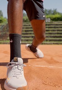Scarpe da tennis grigie con dettagli arancioni, indossate con calze nere a coste, su un campo in terra battuta. Sullo sfondo si vede un terreno livellato con delle scale.
