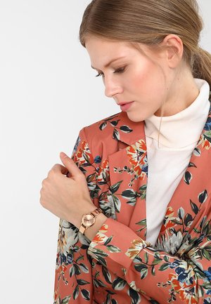 Blazer med blomsterdesign i koralfarve med grønne og blå blade, lagd over en hvid højhalset trøje. Rosaguld ur på handlen, med en rund urskive.
