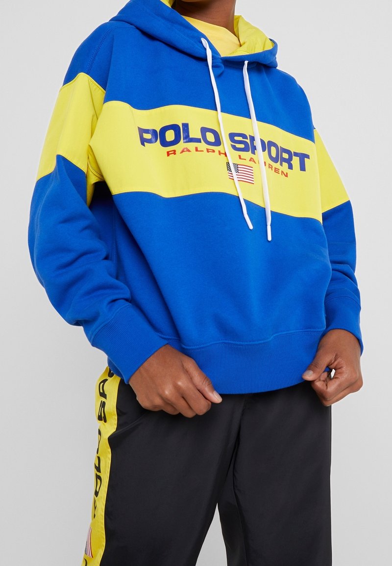 Personne portant un sweat à capuche Polo Sport Ralph Lauren bleu et jaune avec un pantalon noir assorti, avec des bandes latérales jaunes et un logo.