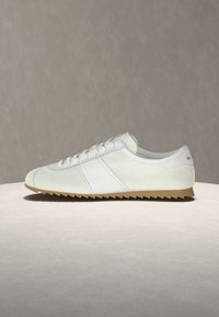 Λευκό sneaker με χαμηλό προφίλ, κορδόνια, σόλα από καουτσούκ και διακριτικές λεπτομέρειες στα πλαϊνά και στη φτέρνα, τοποθετημένο σε φωτεινή επιφάνεια με απλό φόντο.