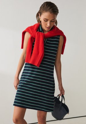 Kvinde iført navyblå og hvide stribede ærmeløse kjole med rød sweater draperet over skuldrene, holder en lille mørk håndtaske.