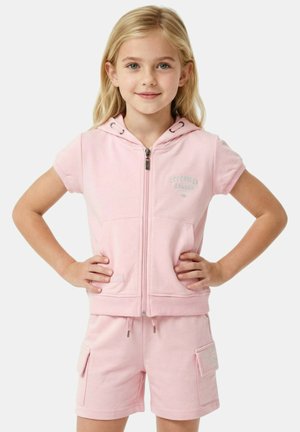 Fille blonde debout, les mains sur les hanches, portant un haut zippé à capuche rose clair et un short cargo assorti.