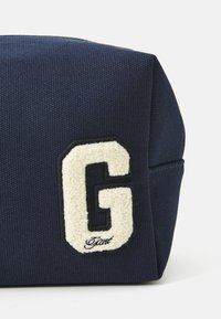 Pochette en tissu bleu marine avec un patch en texte blanc représentant la lettre « G » entourée de noir, et un petit texte en cursive « Gant » en dessous de la lettre.