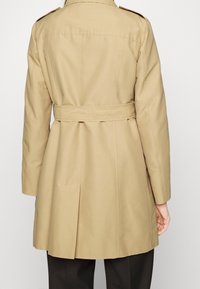 Beżowy trencz z paskiem, szwami tylnymi i wentylacją z tyłu. Posiada naramienniki i dopasowany krój.