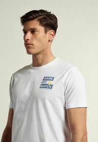 Camiseta de algodón blanca con un estampado gráfico en azul y amarillo que dice "GAME SET MATCH" en la parte izquierda del pecho. Diseñada con mangas cortas y cuello redondo.