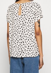 Top a maniche corte in un tessuto leggero con base crema e motivo a macchie nere. Presenta una chiusura a occhio di pernice sul retro.