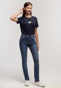 Granatowa koszulka z białym logo, zestawiona z ciemnoniebieskimi dopasowanymi jeansami oraz białymi trampkami. Strój cechuje się klasycznym wzorem i swobodnym stylem.