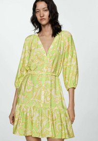 Rochie cu un model verde și galben, cu guler în V, mâneci bufante, închidere cu nasturi și tiv cu volane în straturi. Fabricată dintr-un material ușor.