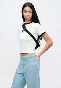 Νεαρή γυναίκα με μακριά μαύρα μαλλιά, που φοράει λευκό κοντό τοπ, ανοιχτό μπλε τζιν παντελόνι και μαύρη τσάντα χιαστί, στέκεται μπροστά σε μια απλή φόντο.