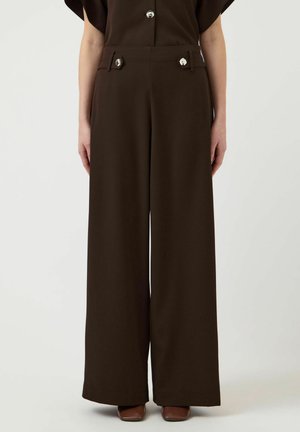 Kvinde iført brune bukser med højtaljede brede ben og dekorative sølvknapper på frontlommerne, kombineret med brune sko med firkantet tå.