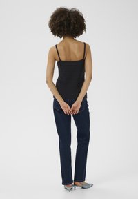 Melna adīta topiņš ar plānām siksnām, savienots ar tumši ziliem džinsiem un gaiši ziliem norādītu pirkstu apaviem, skatīts no aizmugures.