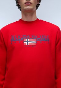 Červená mikina s kulatým výstřihem. Přední design zobrazuje "NAPAPIJRI" v modré barvě a norskou vlajku. Text zahrnuje "SINCE 1987." Bavlněný materiál.