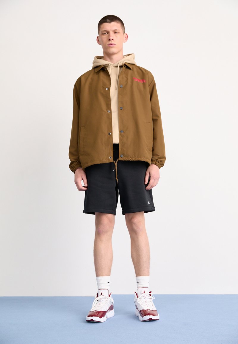 Veste légère marron avec des boutons pression, sweat à capuche beige en dessous, shorts noirs et baskets blanches avec des accents rouges. Fond uni.