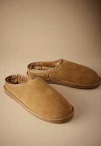 Suede slippers in tan with a soft fur lining, rounded toe, and slip-on design. Features a flexible rubber sole and simple stitching detail.
