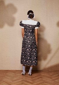 Φλοράλ φόρεμα με φουσκωτά μανίκια, μαύρη βάση και ροζ λουλούδια, λευκό γιακά, φορεμένο με ανοιχτογάλαζες κάλτσες και κρεμ τακούνια, εμφανίζεται από πίσω.