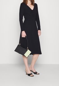 Robe noire à manches longues avec un décolleté en V et des détails froncés, assortie à un sac à main noir structuré et des escarpins noirs ouverts.