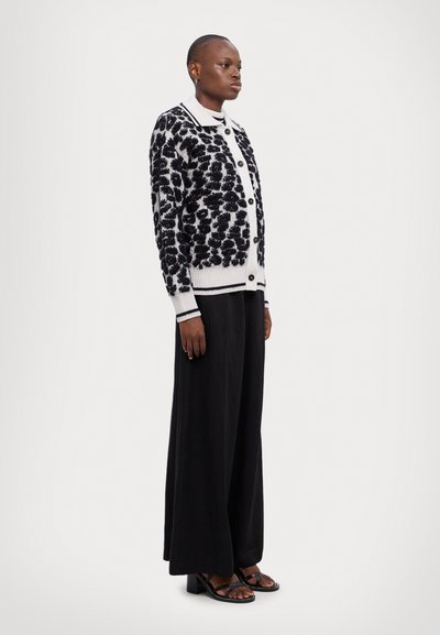 Cardigan noir et blanc avec un motif floral texturé, en maille, à boutonnière, avec des poignets côtelés, associé à un pantalon noir ample et des talons.