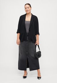 Blazer noir oversize sur un haut gris, associé à une longue jupe en denim noir avec une fente à l'avant. Les accessoires incluent un petit sac à main noir et des talons.