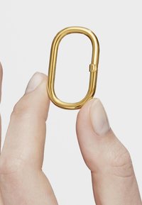 Mano sosteniendo un pequeño mosquetón ovalado dorado entre el pulgar y el índice contra un fondo liso.