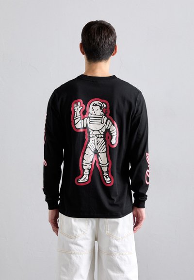 Man facing away wearing black long-sleeve shirt with cartoon astronaut outlined in red on back, paired with white pants.