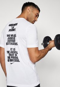 Camiseta blanca de Nike de manga corta con texto negro en la parte trasera que incluye citas motivacionales y el logo de Nike. El modelo sostiene un mancuerna.