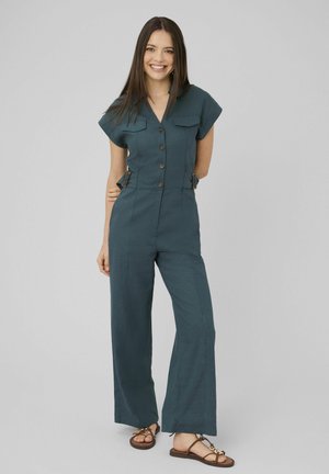 Femme souriante aux longs cheveux foncés, vêtue d'une combinaison teal boutonnée et de sandales plates marron, debout devant un fond clair uni.