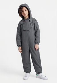 Kind gekleed in een grijs jumpsuit met capuchon en een ritssluiting aan de voorkant, en witte sneakers, staand tegen een effen lichte achtergrond.